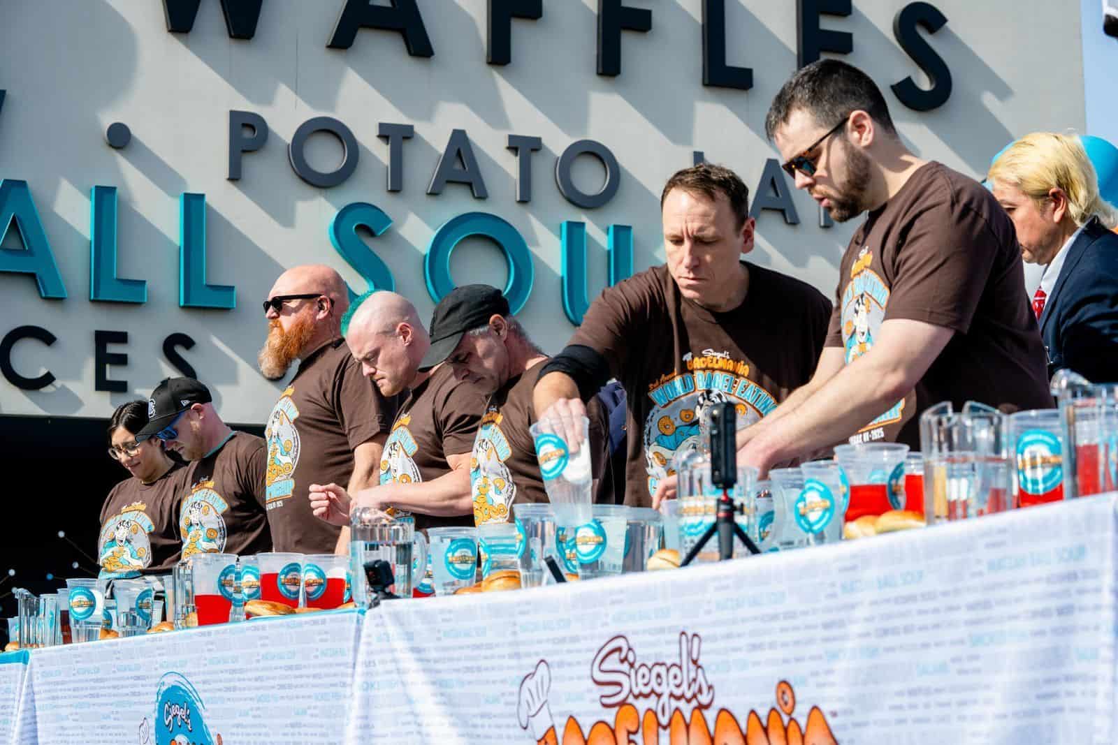 Contestants lined up at tables during a bagel eating competition, preparing and eating under a large event banner.